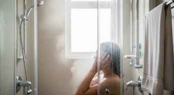 Vista lateral de una persona duchándose con vapor en una cabina de cristal, con el cabezal de la ducha y una toalla limpia colgando a la derecha.