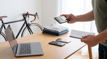 Hombre sostiene un smartphone y una libreta. Sobre mesa de madera: laptop abierta, dos móviles, ropa doblada. Bicicleta en segundo plano.