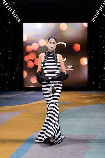 Modelo en pasarela con vestido de rayas blancas y negras, cuello negro brillante y guantes voluminosos, sosteniendo un bolso. Fondo oscuro con pantalla