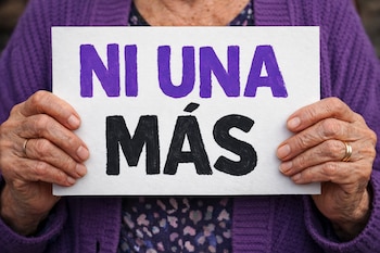 Primer plano de manos de una mujer con anillos, sosteniendo un cartel blanco con la frase Ni Una Más en letras moradas y negras, frente a un chaleco morado.