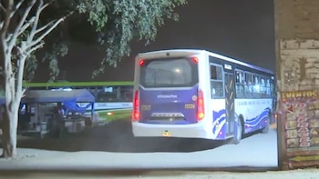Un autobús de color morado y blanco circula de noche cerca de un árbol y un muro con afiches publicitarios