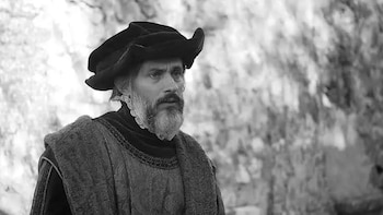 Primer plano de un hombre barbudo con bigote, sombrero y vestimenta de época con gorguera, en blanco y negro, con fondo borroso