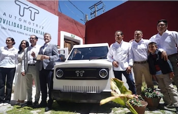 Grupo de personas sonríe y posa frente a un auto eléctrico blanco con el logo 'TT' y un banner de 'Movilidad Sustentable' en un evento al aire libre