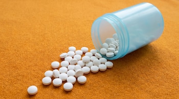 Un frasco de plástico celeste, sin etiquetas, está volcado sobre una superficie naranja texturizada, con numerosas pastillas blancas redondas desparramadas a su lado.