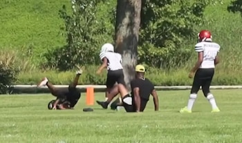 Tres jóvenes con cascos de fútbol americano y un adulto con gorra realizan ejercicios en un campo de hierba