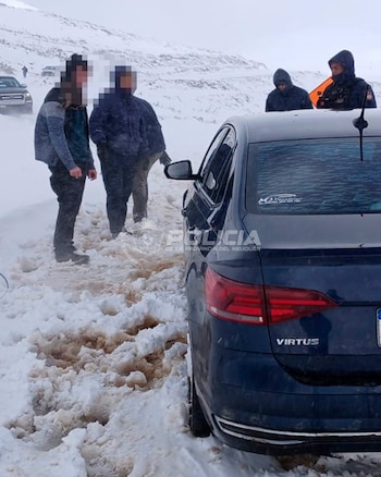 Varias personas abrigadas y efectivos policiales permanecen junto a un automóvil azul sobre un camino cubierto de abundante nieve