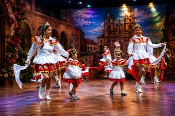 Mujeres y niñas con trajes blancos y rojos, pañuelos y coronas bailan sobre un escenario de madera con un telón de fondo urbano colonial