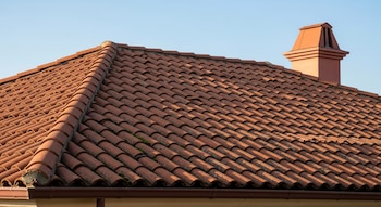 Vista de un techo de tejas de arcilla curvas de color rojizo-marrón con una chimenea a la derecha, bajo un cielo azul claro. Un canalón oscuro corre a lo largo del borde inferior del tejado.