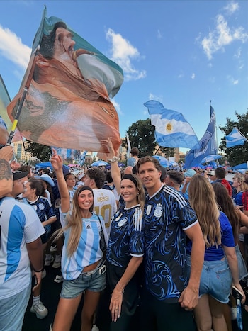 Multitud de personas celebra con banderas de Argentina y camisetas de fútbol. Se distinguen una bandera grande con retrato y el cielo azul