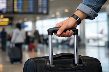 Primer plano de la mano de una persona sujetando el asa de una maleta de ruedas negra en una terminal de aeropuerto, con otras personas al fondo.