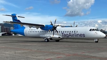 Avión blanco y azul de Tag Airlines con hélices visibles, estacionado en una pista de asfalto bajo un cielo parcialmente nublado. Se ven edificios al fondo