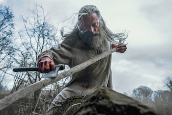 Hombre anciano de cabello y barba largos con túnica. Sostiene un hacha con la mano derecha, manchada de sangre. Un tronco y árboles secos al fondo