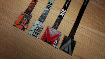 Cuatro medallas de colores naranja, celeste, rojo y gris oscuro, con sus cintas, dispuestas en diagonal sobre una superficie de madera. Incluyen textos y diseños