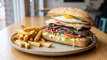 Sándwich de lomito completo con carne de lomo, jamón, queso, huevo frito, tomate, lechuga, cebolla y papas fritas en un plato sobre una mesa de madera.