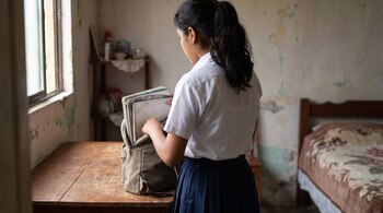 Una joven con uniforme escolar guarda cuadernos en una mochila sobre una mesa de madera dentro de una habitación.