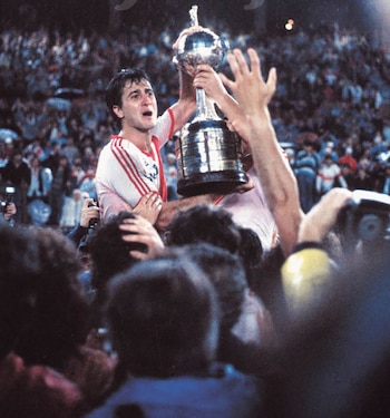 Un hombre con camiseta deportiva blanca y roja levanta un trofeo metálico rodeado por una multitud de personas en un estadio