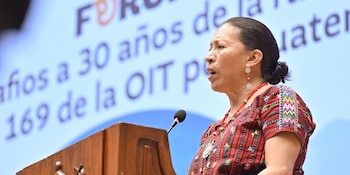 Mujer de cabello oscuro con camisa bordada y aretes redondos habla ante un micrófono en podio de madera. Pantalla azul con texto detrás