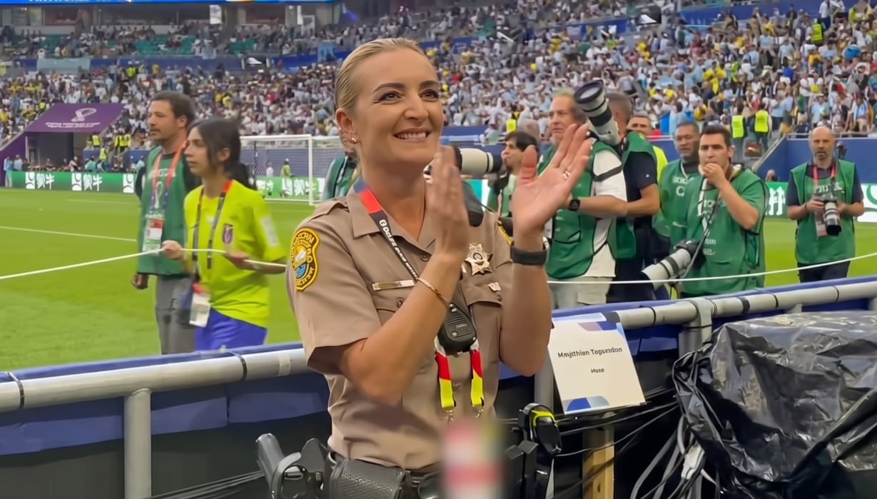 Una oficial de seguridad con uniforme color caqui y distintivos en el pecho aplaude sonriente frente a una multitud en un estadio de fútbol (Captura de video)