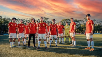 Once jóvenes futbolistas con uniformes rojos y blancos, y un portero con uniforme amarillo, posan en un campo de fútbol al atardecer con un cielo naranja