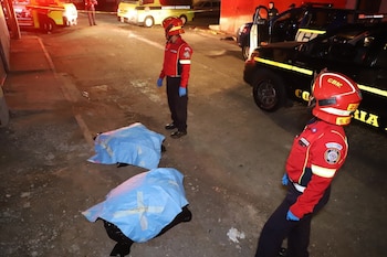 Dos cuerpos cubiertos con sábanas azules en el suelo, dos bomberos con uniformes rojos, cascos y guantes, ambulancias, un vehículo policial oscuro, luces de emergencia y una calle de noche
