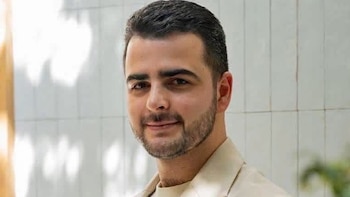 Primer plano de un hombre con cabello oscuro corto y barba, viste una camisa clara con cuello, mira al frente y sonríe ligeramente. Fondo claro y difuso