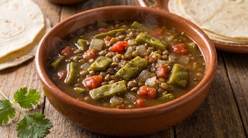 Un plato de barro con sopa de lentejas, trozos de nopal y jitomate, acompañado de tortillas de maíz sobre una mesa de madera.