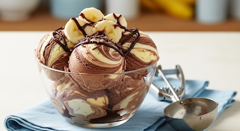 Un tazón de vidrio transparente lleno de bolas de helado marmoleado de chocolate y plátano, decorado con rodajas de plátano, sirope y virutas de chocolate.