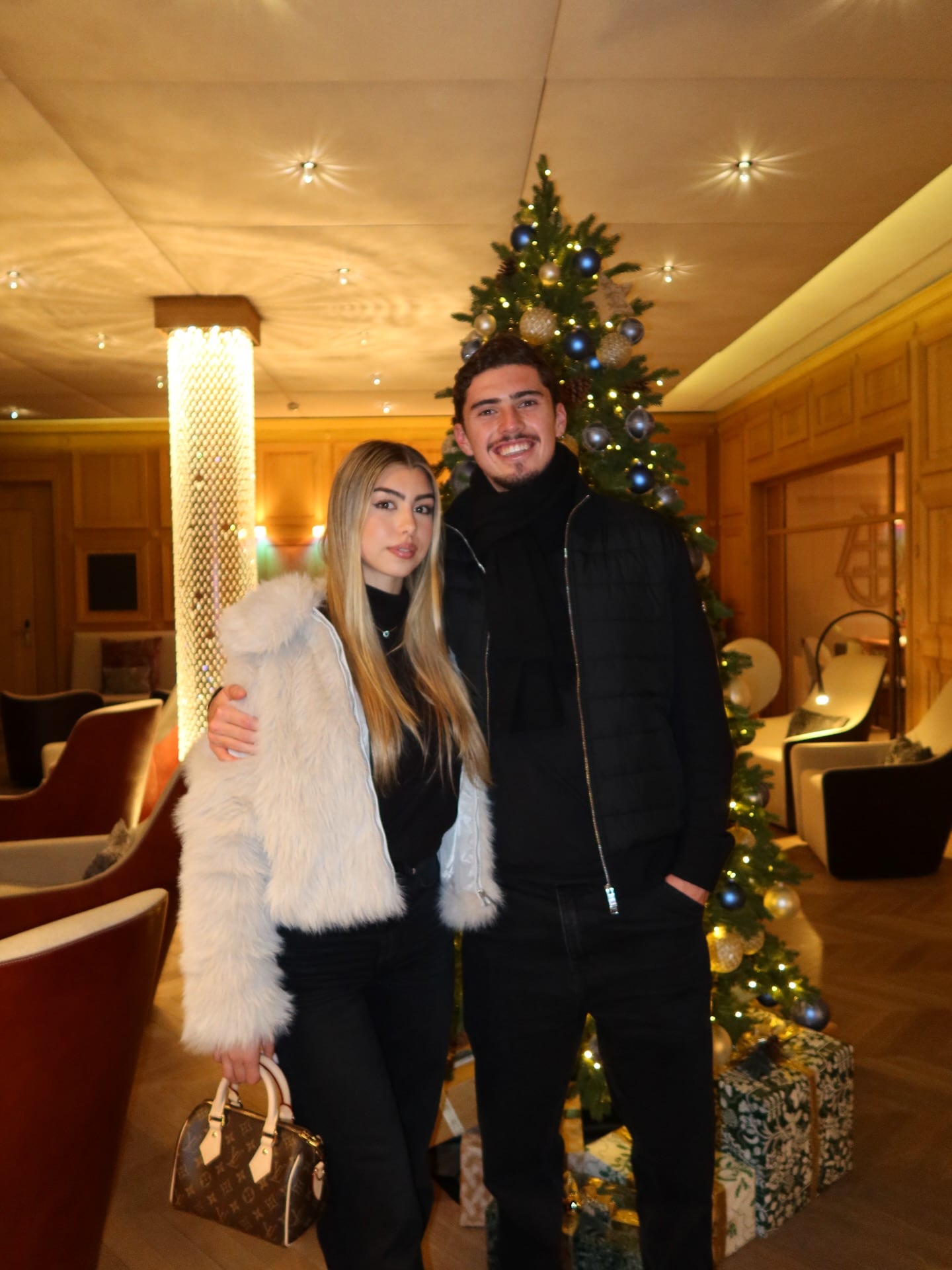 Joaquín Panichelli y Giuliana Giramonti posan de pie junto a un árbol de Navidad decorado con luces y esferas azules y doradas en un ambiente interior.