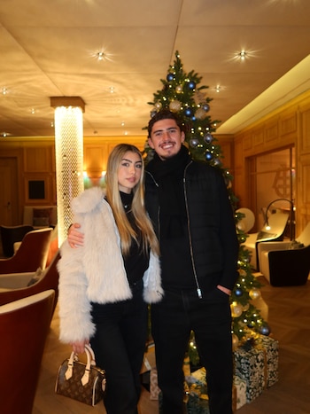 Hombre y mujer posan de pie junto a un árbol de Navidad iluminado con esferas azules y doradas, con regalos envueltos en su base, en un interior con luz tenue