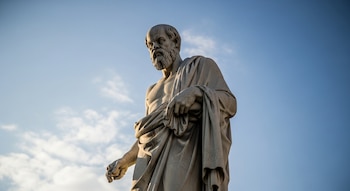 Estatua de Sócrates de mármol, en ángulo contrapicado, con un cielo azul y nubes blancas de fondo.
