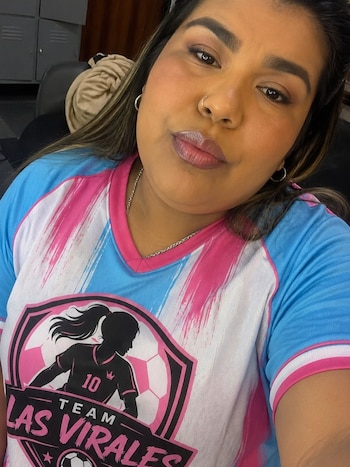 Primer plano de una mujer con piel trigueña y cabello oscuro, usando una camiseta deportiva rosa, blanca y azul con un logo de fútbol femenino y un collar plateado