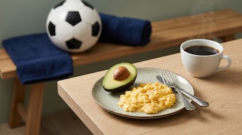 Plato con huevos revueltos, medio aguacate con semilla, café humeante y cubiertos en mesa de madera clara. Al fondo, un balón de fútbol y una toalla azul.