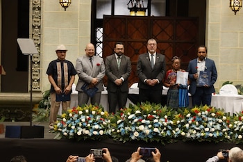 Seis personas en un escenario. Documentos y una flor en las manos de algunos participantes. Arreglos florales. Pared de piedra con puertas y lámparas. Manos con teléfonos móviles