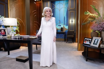 Una mujer de cabello rubio con vestido blanco largo está de pie en un estudio de televisión. Un escritorio negro con lámpara y adornos la acompaña
