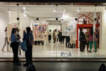 Personas contemplan obras textiles bordadas colgadas en una sala iluminada vista desde la calle a través de un ventanal