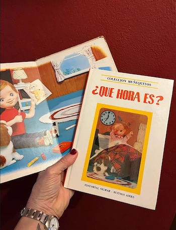 Una mano sostiene un libro infantil blanco titulado "¿Qué hora es?". En el fondo, un libro abierto con ilustraciones de niños y perros sobre una superficie roja
