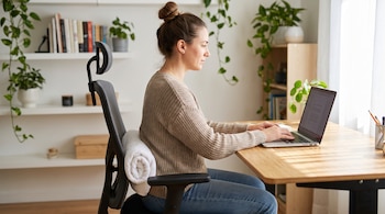 Mujer sentada de perfil frente a un portátil en un escritorio de madera, con una toalla blanca enrollada como soporte lumbar en una silla de oficina.