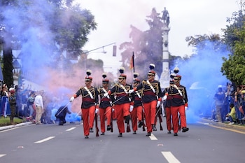 Un grupo de soldados uniformados marcha con sables en una calle, rodeados de humo azul y rosa. Hay público a los costados y vegetación al fondo