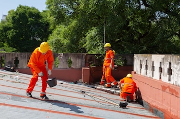 Tres trabajadores de la construcción con uniformes naranjas y cascos amarillos trabajan en un tejado metálico. Se ven herramientas esparcidas. Árboles verdes de fondo
