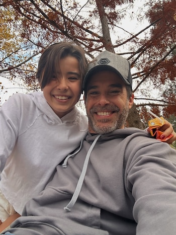 Hombre con gorra gris y niña con capucha blanca sonríen en un primer plano. La niña sostiene un envase de jugo. De fondo, ramas de árbol con hojas de otoño