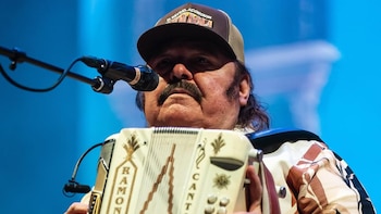 Primer plano de Ramón Ayala con un sombrero marrón, bigote, tocando un acordeón blanco personalizado en un escenario con un micrófono