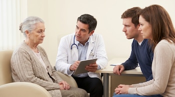 Una anciana de cabello gris sentada frente a un médico con tableta y dos familiares. Todos están sentados en una oficina con fondo claro.