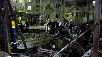 Personal de rescate trabaja de noche entre automóviles destruidos y escombros frente a un edificio con ventanas rotas