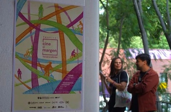 Un cartel blanco con ilustraciones coloridas y texto sobre un festival de cine en una pared blanca. Dos personas conversan frente a árboles