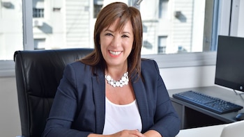 Retrato de la abogada Mariana Barbitta sonriendo a cámara, sentada en una silla de oficina negra. Viste un saco azul marino y un collar blanco