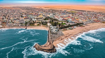 Vista aérea de una ciudad costera con edificios, carreteras, vehículos, un muelle rocoso, una playa arenosa, el océano con olas y un desierto de arena al fondo.