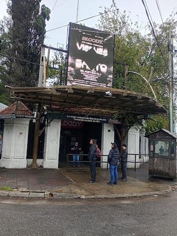 Vista frontal de la entrada de un edificio con un cartel LED grande. Varios hombres parados en la acera mojada. Un cobertizo de madera a la derecha y árboles al fondo