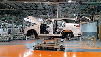 Vista interior de una fábrica de automóviles con el chasis blanco de un SUV en la línea de montaje, rodeado de maquinaria industrial y otros vehículos en construcción