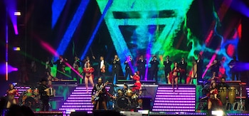 Músicos vestidos de traje con instrumentos de viento y cuerda actúan en un escenario iluminado junto a bailarinas con trajes rojos