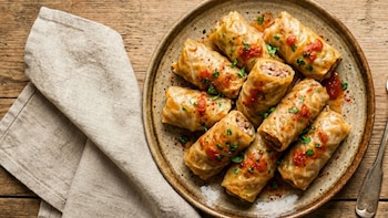 Plato rústico con rollos de col rellenos sobre una mesa de madera, acompañado de una servilleta de lino, salsa de tomate, perejil fresco y un tenedor